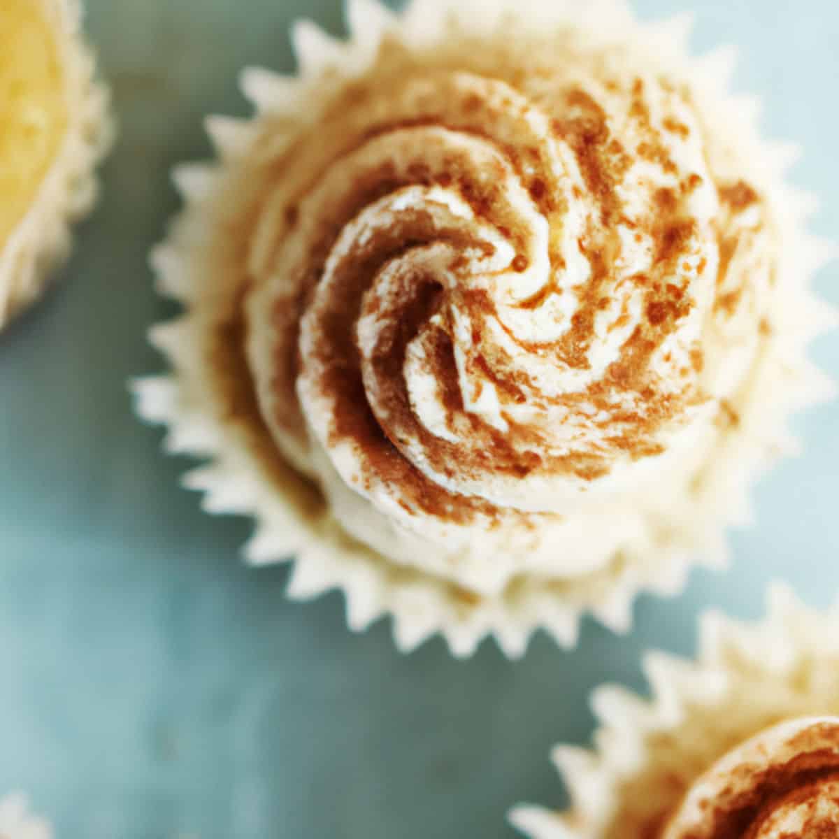 Delicious cupcake with swirled vanilla frosting sprinkled with cocoa powder, in a white paper cup, on a pastel background.